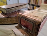 A rustic and worn index card catalog is accompanied by two old, thick books with noticeable aging and distressed covers. The background features a blurred array of books on shelves, enhancing the vintage and library-like ambiance of the scene.