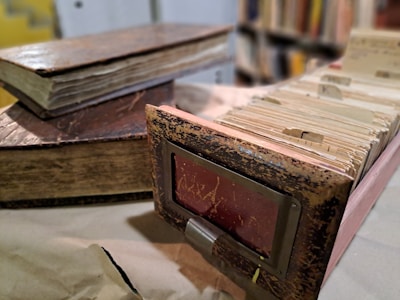 A rustic and worn index card catalog is accompanied by two old, thick books with noticeable aging and distressed covers. The background features a blurred array of books on shelves, enhancing the vintage and library-like ambiance of the scene.