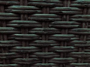 A close-up view of a woven pattern with a textured, grid-like structure. The material appears to be a dark, possibly synthetic fiber that crisscrosses to form a rigid surface. Each strand has a slightly glossy finish, adding depth and dimension to the pattern.