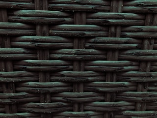 A close-up view of a woven pattern with a textured, grid-like structure. The material appears to be a dark, possibly synthetic fiber that crisscrosses to form a rigid surface. Each strand has a slightly glossy finish, adding depth and dimension to the pattern.
