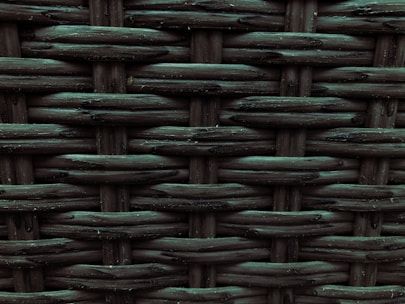 A close-up view of a woven pattern with a textured, grid-like structure. The material appears to be a dark, possibly synthetic fiber that crisscrosses to form a rigid surface. Each strand has a slightly glossy finish, adding depth and dimension to the pattern.