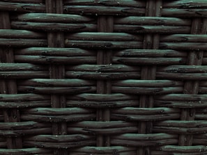 A close-up view of a woven pattern with a textured, grid-like structure. The material appears to be a dark, possibly synthetic fiber that crisscrosses to form a rigid surface. Each strand has a slightly glossy finish, adding depth and dimension to the pattern.