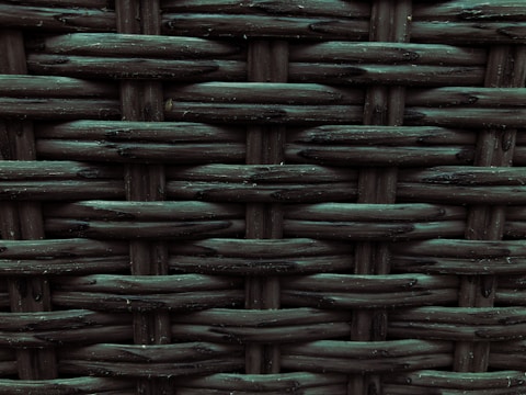 A close-up view of a woven pattern with a textured, grid-like structure. The material appears to be a dark, possibly synthetic fiber that crisscrosses to form a rigid surface. Each strand has a slightly glossy finish, adding depth and dimension to the pattern.