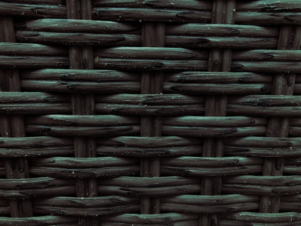 A close-up view of a woven pattern with a textured, grid-like structure. The material appears to be a dark, possibly synthetic fiber that crisscrosses to form a rigid surface. Each strand has a slightly glossy finish, adding depth and dimension to the pattern.