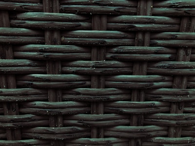 A close-up view of a woven pattern with a textured, grid-like structure. The material appears to be a dark, possibly synthetic fiber that crisscrosses to form a rigid surface. Each strand has a slightly glossy finish, adding depth and dimension to the pattern.