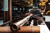 A traditional Japanese water basin with long wooden ladles placed across the edge. The background consists of blurred architectural and natural elements, suggesting a serene and historic setting.