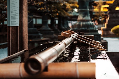 A traditional Japanese water basin with long wooden ladles placed across the edge. The background consists of blurred architectural and natural elements, suggesting a serene and historic setting.