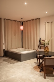A bathroom with a modern bathtub surrounded by beige curtains. Above the tub hangs a single red pendant light, creating a cozy ambiance. The floor is tiled in a light marble design, and there is a small round table with chairs next to the tub. A tall vase with flowers adorns the table, adding a touch of elegance to the space.