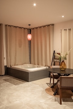 A bathroom with a modern bathtub surrounded by beige curtains. Above the tub hangs a single red pendant light, creating a cozy ambiance. The floor is tiled in a light marble design, and there is a small round table with chairs next to the tub. A tall vase with flowers adorns the table, adding a touch of elegance to the space.