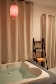 A cozy bathroom setting features a white bathtub filled with water, surrounded by cream-colored curtains. A red pendant light hangs above, casting a warm glow. A sleek black shelf holds rolled yellow towels and potted plants.