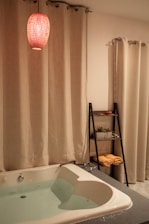 A cozy bathroom setting features a white bathtub filled with water, surrounded by cream-colored curtains. A red pendant light hangs above, casting a warm glow. A sleek black shelf holds rolled yellow towels and potted plants.