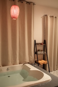 A cozy bathroom setting features a white bathtub filled with water, surrounded by cream-colored curtains. A red pendant light hangs above, casting a warm glow. A sleek black shelf holds rolled yellow towels and potted plants.