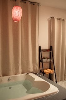 A cozy bathroom setting features a white bathtub filled with water, surrounded by cream-colored curtains. A red pendant light hangs above, casting a warm glow. A sleek black shelf holds rolled yellow towels and potted plants.