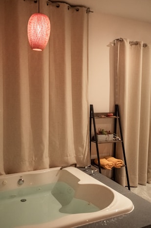 A cozy bathroom setting features a white bathtub filled with water, surrounded by cream-colored curtains. A red pendant light hangs above, casting a warm glow. A sleek black shelf holds rolled yellow towels and potted plants.