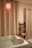 A cozy bathroom setting features a white bathtub filled with water, surrounded by cream-colored curtains. A red pendant light hangs above, casting a warm glow. A sleek black shelf holds rolled yellow towels and potted plants.