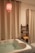 A cozy bathroom setting features a white bathtub filled with water, surrounded by cream-colored curtains. A red pendant light hangs above, casting a warm glow. A sleek black shelf holds rolled yellow towels and potted plants.