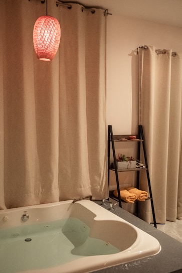 A cozy bathroom setting features a white bathtub filled with water, surrounded by cream-colored curtains. A red pendant light hangs above, casting a warm glow. A sleek black shelf holds rolled yellow towels and potted plants.