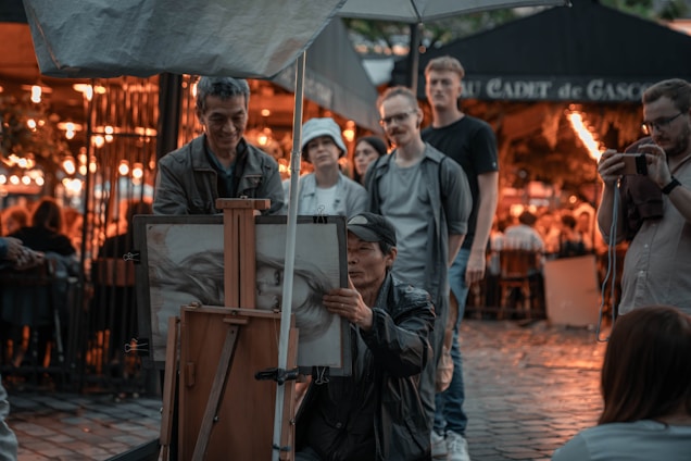 A street artist is sketching a portrait on an easel while a small group of people observe the work. The setting appears to be an outdoor area, possibly a market or café, with warm lights illuminating the background.