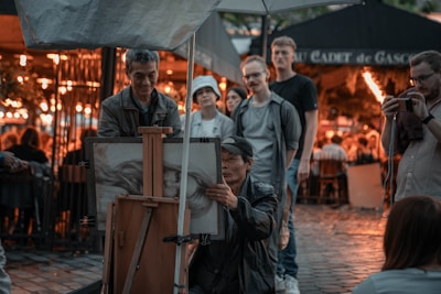 A street artist is sketching a portrait on an easel while a small group of people observe the work. The setting appears to be an outdoor area, possibly a market or café, with warm lights illuminating the background.