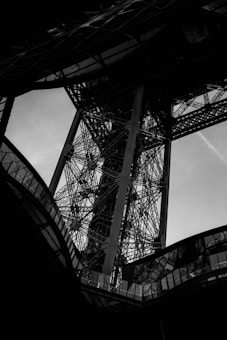 A black and white close-up view of a complex metal structure with intersecting beams and girders. The image shows intricate ironwork with an abstract and geometric appearance, creating a dramatic composition.