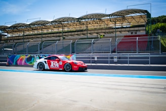 A red and white racing car is positioned on a racetrack, adjacent to a grandstand with empty seats. The grandstand is covered with a green roof structure, and fencing separates the track and the stands. Bright murals decorate the barriers behind the car, and the scene is illuminated by a clear blue sky.