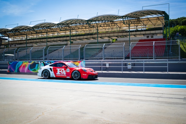 A red and white racing car is positioned on a racetrack, adjacent to a grandstand with empty seats. The grandstand is covered with a green roof structure, and fencing separates the track and the stands. Bright murals decorate the barriers behind the car, and the scene is illuminated by a clear blue sky.