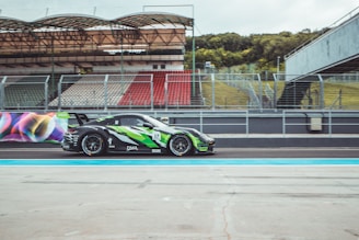 A race car with a sleek design featuring green and black colors speeds along a racing track. The car is adorned with various sponsor logos and the number 17 is prominently displayed. The background includes an empty grandstand with red and white seats and a green grassy area.