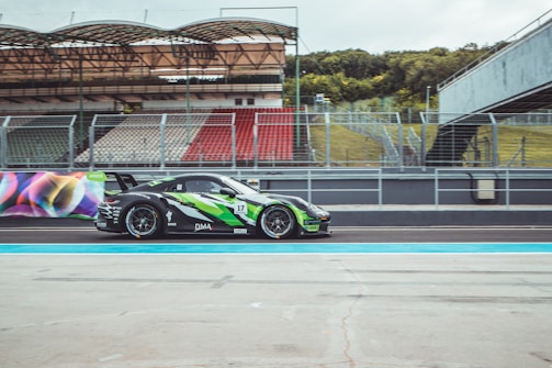 A race car with a sleek design featuring green and black colors speeds along a racing track. The car is adorned with various sponsor logos and the number 17 is prominently displayed. The background includes an empty grandstand with red and white seats and a green grassy area.