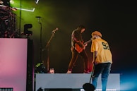 A stage performance with two musicians. One is playing an electric guitar while the other stands nearby, holding a microphone and wearing a yellow jersey with the number 92. Stage lights create a vibrant atmosphere with green and magenta hues.