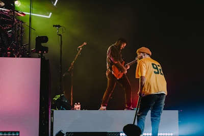 A stage performance with two musicians. One is playing an electric guitar while the other stands nearby, holding a microphone and wearing a yellow jersey with the number 92. Stage lights create a vibrant atmosphere with green and magenta hues.
