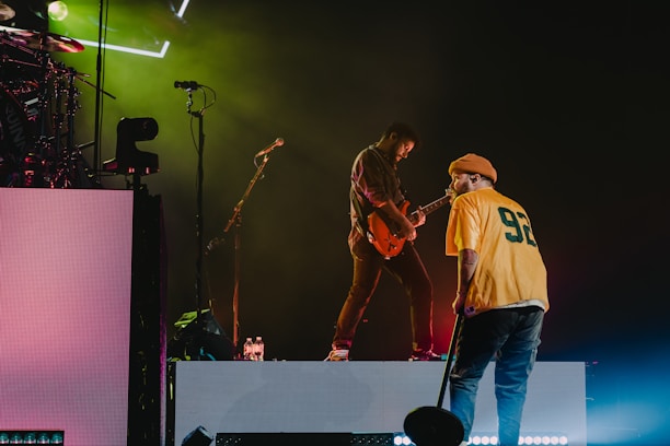 A stage performance with two musicians. One is playing an electric guitar while the other stands nearby, holding a microphone and wearing a yellow jersey with the number 92. Stage lights create a vibrant atmosphere with green and magenta hues.