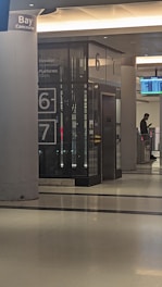 An interior of a transit station featuring an elevator with platform numbers 6 and 7 displayed prominently. There is a digital screen displaying train information and a silhouette of a person standing nearby, possibly checking their phone. The environment is clean with modern architectural elements.