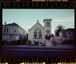 A quaint, historic building with large, ornate stained glass windows is flanked by two residential homes. The building is painted in pastel tones with an arched roof and a small tower on the right-hand side. A red awning with 'BELLROSE' written on it covers the entrance. Palm trees and shrubs are planted around the base. The sky is clear, and the photo has a film reel border indicating it was taken on Kodak Portra 400 film.