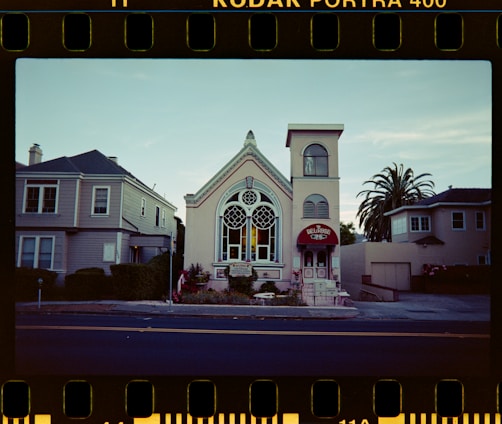 A quaint, historic building with large, ornate stained glass windows is flanked by two residential homes. The building is painted in pastel tones with an arched roof and a small tower on the right-hand side. A red awning with 'BELLROSE' written on it covers the entrance. Palm trees and shrubs are planted around the base. The sky is clear, and the photo has a film reel border indicating it was taken on Kodak Portra 400 film.