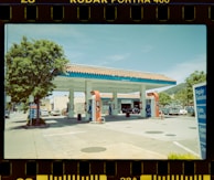 A vintage-style photograph of a gas station featuring pastel tones and film borders. The station has two gas pumps under a canopy with a tiled roof, and several vehicles are parked nearby. There is clear signage indicating fuel types, with some trees and a mountain visible in the background.