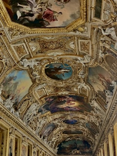 An ornate and intricately decorated ceiling featuring detailed paintings and elaborate gold embellishments. The artwork includes classical figures and mythological scenes, framed by opulent gilded moldings and surrounded by sculpted cherubs and floral motifs.