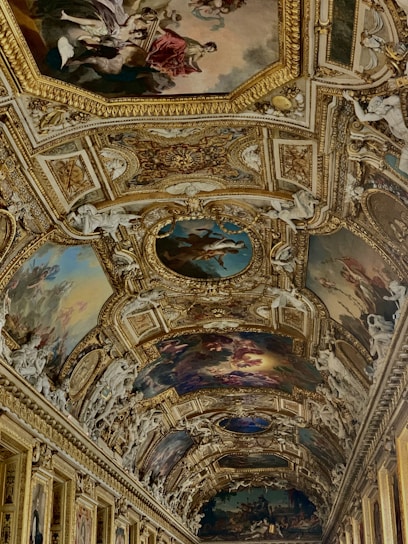 An ornate and intricately decorated ceiling featuring detailed paintings and elaborate gold embellishments. The artwork includes classical figures and mythological scenes, framed by opulent gilded moldings and surrounded by sculpted cherubs and floral motifs.