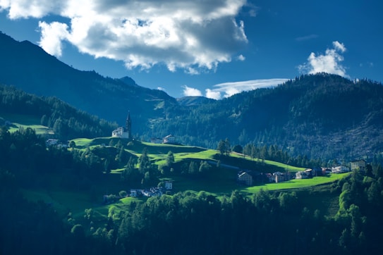 A picturesque mountainous landscape with lush green hills, scattered houses, and a prominent church with a tall steeple. The sky is partly cloudy, and the scene is enveloped in a serene atmosphere. Sunlight highlights the vibrant green pastures, while the surrounding mountains provide a dark, contrasting backdrop.