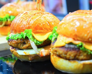 Close-up view of two juicy burgers with sesame seed buns, crisp lettuce, melted cheese slices, and a well-cooked meat patty. The burgers are shown in vibrant colors, emphasizing the fresh ingredients.