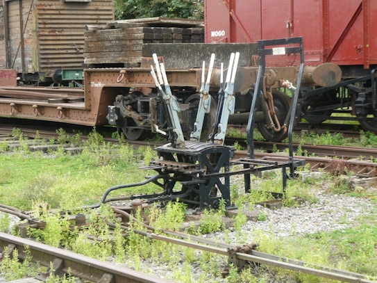 An old railway yard with a set of manual lever switches used for changing tracks. The area is surrounded by overgrown vegetation, and several cargo train wagons are visible in the background.