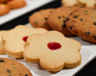 A close-up view of a selection of cookies placed on a white surface. There are flower-shaped cookies with a red center alongside round cookies with chocolate chips.