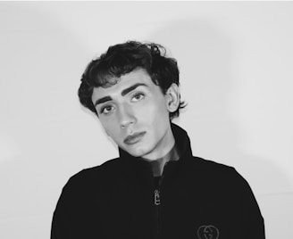 A black and white portrait of a person with short, dark hair and prominent eyebrows, wearing a dark jacket with an emblem on the chest. The backdrop is a plain light color, and the expression is neutral and introspective.