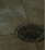 A metal drain cover is set into a tiled floor, with water droplets scattered around. The tiles are a beige color, and the drain has a grid-like pattern.