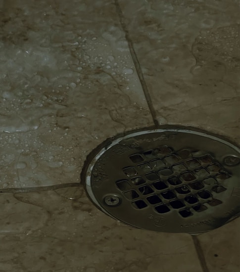 A metal drain cover is set into a tiled floor, with water droplets scattered around. The tiles are a beige color, and the drain has a grid-like pattern.