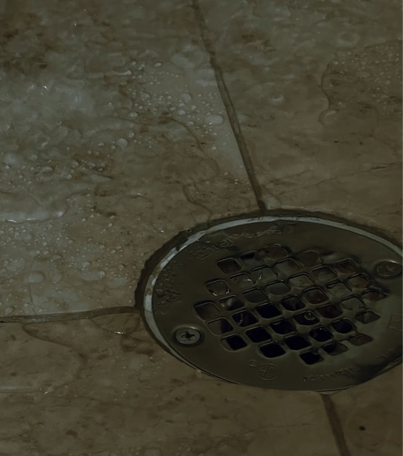 A metal drain cover is set into a tiled floor, with water droplets scattered around. The tiles are a beige color, and the drain has a grid-like pattern.