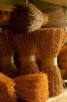 Bundles of closely packed, dried broom-like fibers tied together at the base are arranged in an orderly fashion. The texture is rough and the lighting casts a warm, golden hue on the natural materials, emphasizing their earthy and rustic quality.