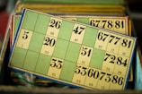 A collection of vintage bingo cards is stacked in a box. The cards have a retro design with numbers printed in bold, black font on a green background. The edges of the cards are bordered with blue.