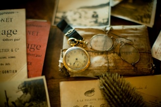 An assortment of vintage items including a pocket watch with a gold chain, a pair of round spectacles, and various old papers and booklets are displayed on a wooden surface. The scene evokes a sense of nostalgia and historical intrigue.