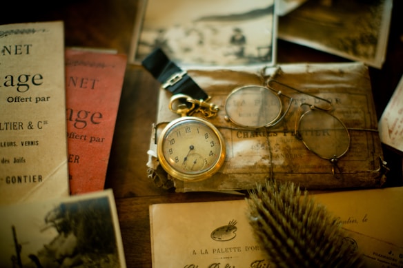 An assortment of vintage items including a pocket watch with a gold chain, a pair of round spectacles, and various old papers and booklets are displayed on a wooden surface. The scene evokes a sense of nostalgia and historical intrigue.