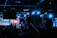Silhouettes of camera operators and equipment set up in a dimly lit venue, illuminated by blue stage lights. A large screen displays colorful graphics in the background.